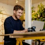 カメラとノートパソコンをテーブルの上に置いて作業をしている男性の画像