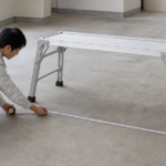 作業台を設置する床を作業者がメジャーで測る場面