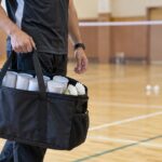 体育館で複数のシャトル筒が入ったバッグを運ぶコーチ