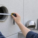 外壁の丸い換気口をメジャーで測り、換気フードを選んでいる様子