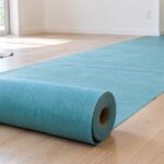 室内の木製床に青緑色の床養生用ロールマットを広げた様子