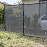 住宅と車の前に飛散防護ネットを置いて草刈りをする作業者