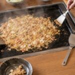 卓上の鉄板でもんじゃ焼きを囲み、金属・樹脂・竹のヘラを並べた様子