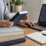 複数の本を手に取り、ノートパソコンの開発画面と見比べている様子